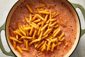 Adding dry penne pasta directly into the creamy spicy vodka sauce base inside a large pot.