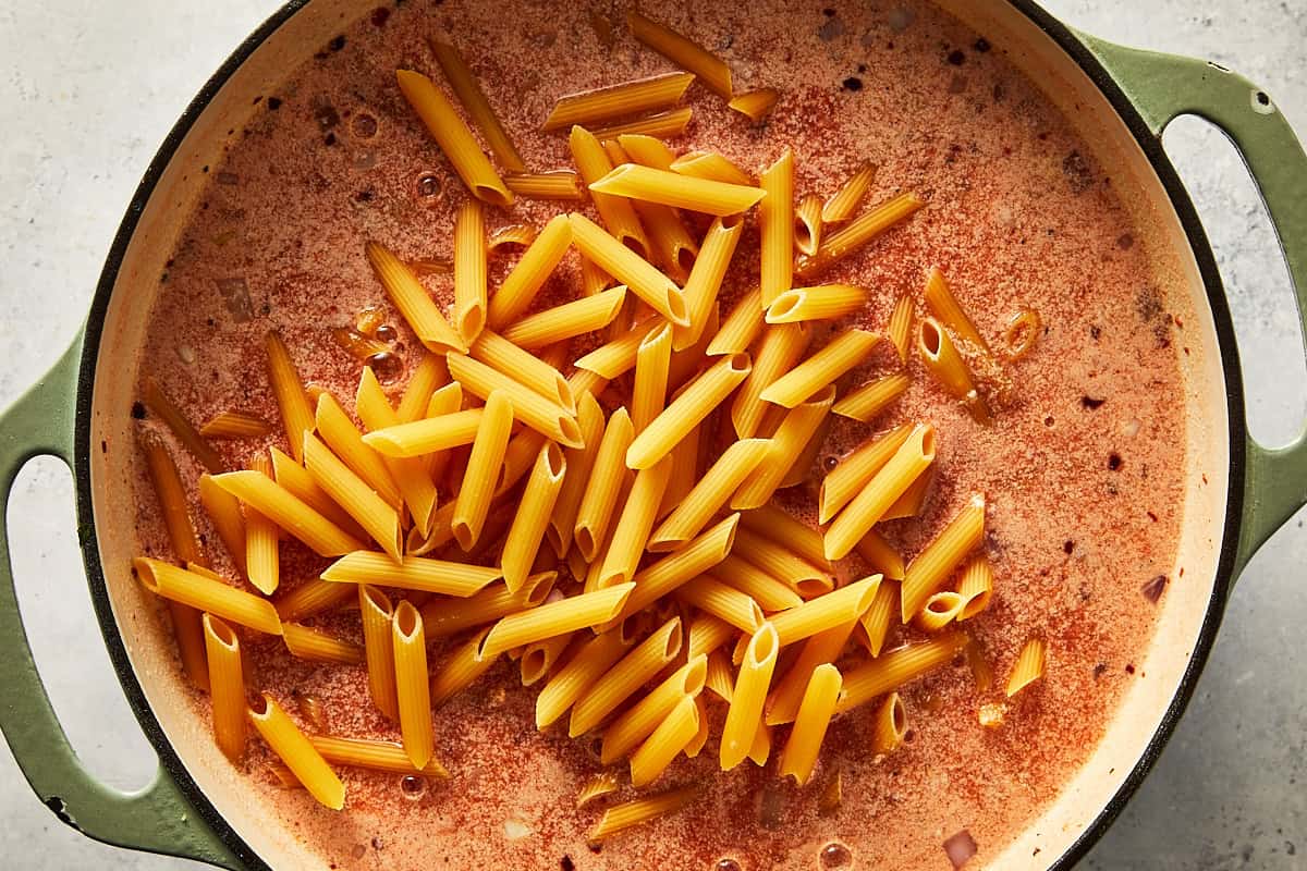 Adding dry penne pasta directly into the creamy spicy vodka sauce base inside a large pot.