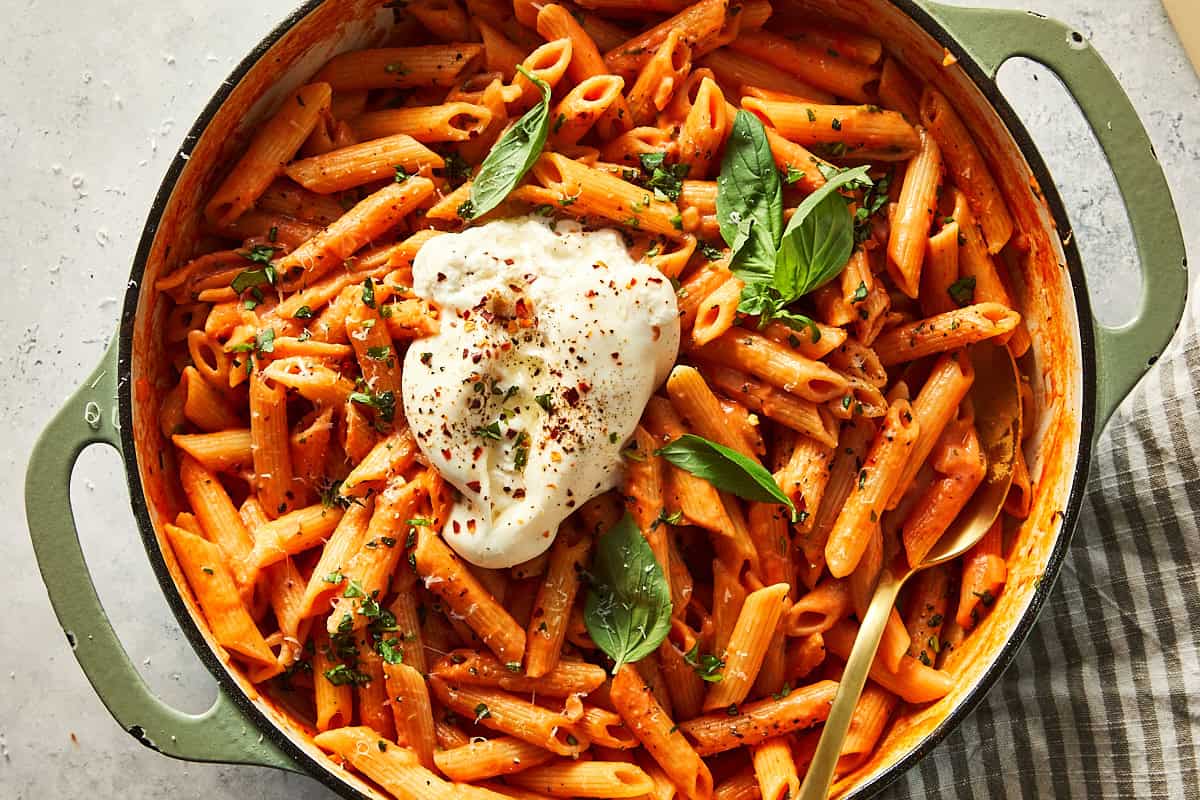 A pot filled with one-pot spicy vodka penne pasta, topped with a torn fresh burrata, fresh basil leaves, and red chili flakes.