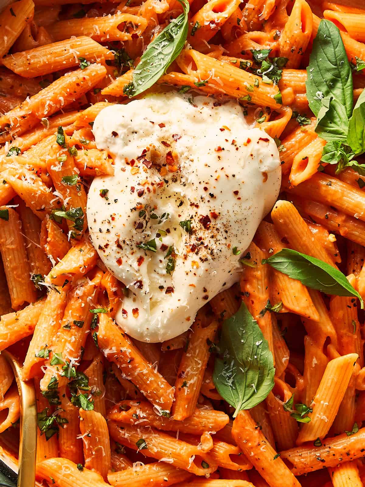 Close-up of one-pot spicy vodka pasta made with penne, topped with a broken ball of fresh burrata, fresh basil leaves, grated cheese, and red chili flakes.