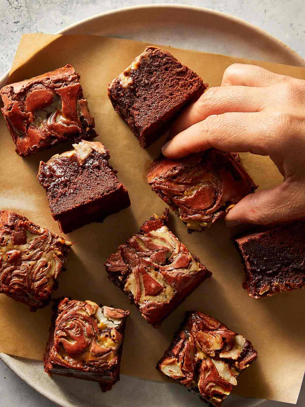 A top-down view of sliced red velvet brownies made from scratch, resting on parchment paper. A hand is reaching to grab a square piece, highlighting the beautiful white chocolate wave pattern on top.