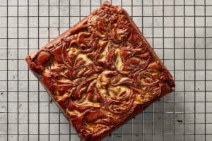 A full, uncut square block of baked homemade red velvet brownie cooling on a wire rack, showcasing the intricate melted white chocolate swirl across the surface.