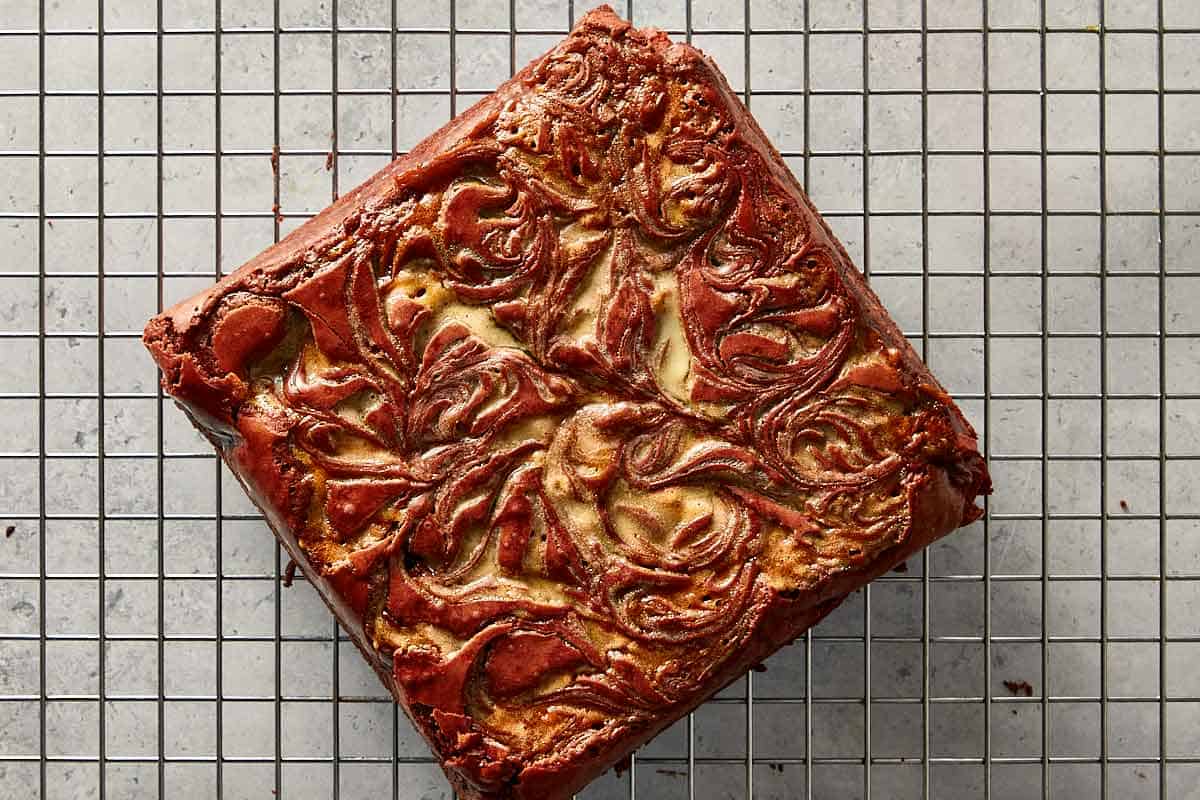 A full, uncut square block of baked homemade red velvet brownie cooling on a wire rack, showcasing the intricate melted white chocolate swirl across the surface.