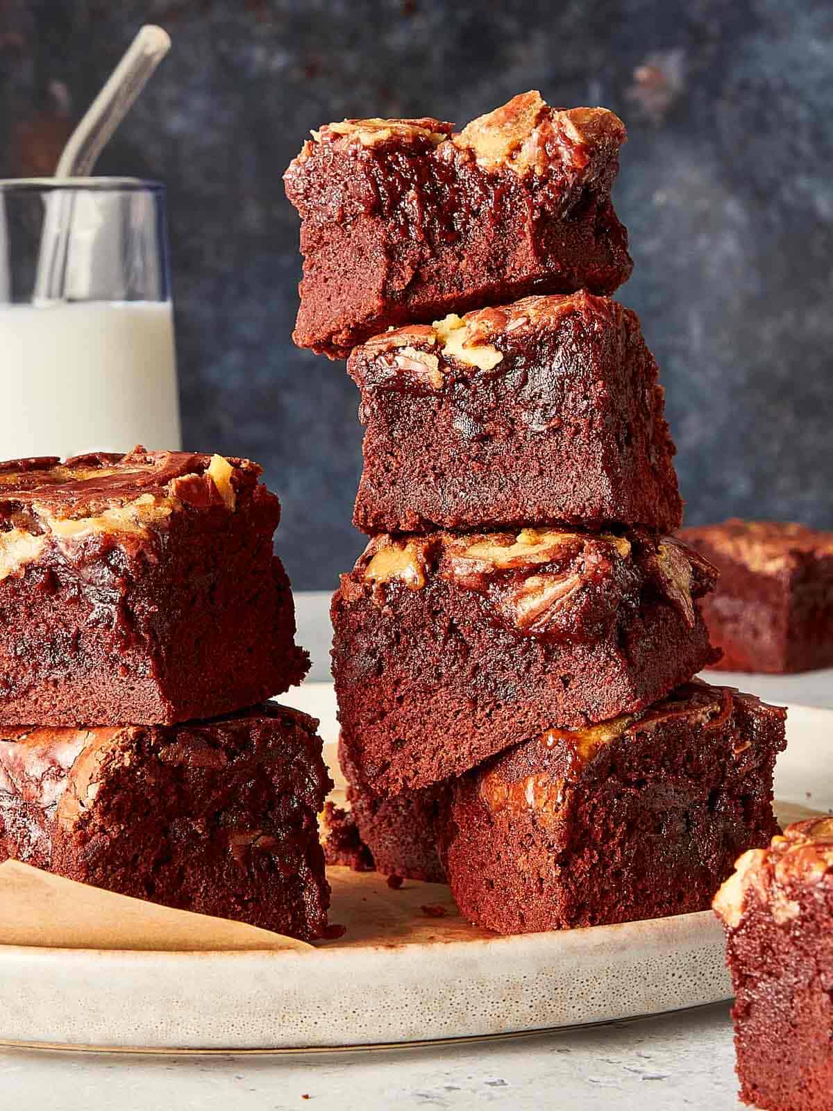 A tall stack of four thick, fudgy homemade red velvet brownies showing their dense texture and white chocolate swirled tops, with a glass of milk in the background.