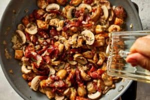 adding wine to a pan with chopped herbs, shallot, grated garlic, and sliced mushrooms