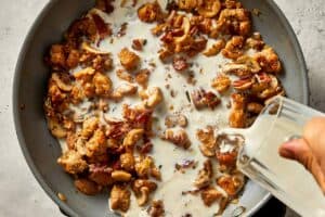 adding milk to a pan with chopped herbs, shallot, grated garlic, and sliced mushrooms