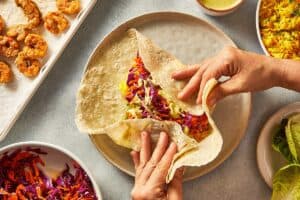 Hands folding a large tortilla over shrimp, rice, slaw, and creamy salsa verde.