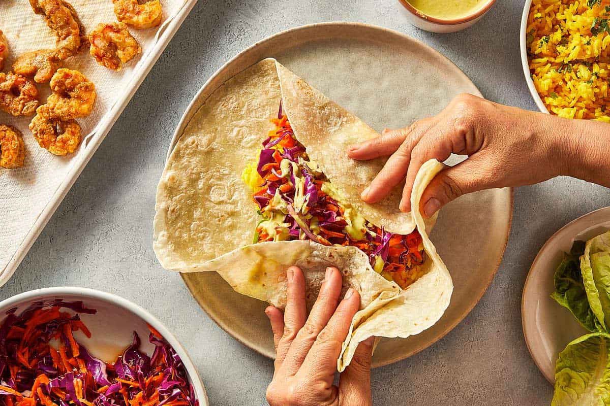 Hands folding a large tortilla over shrimp, rice, slaw, and creamy salsa verde.