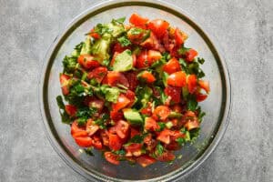 A clear glass bowl filled with fresh diced tomatoes, cubed avocado, chopped cilantro, and jalapenos mixed together.