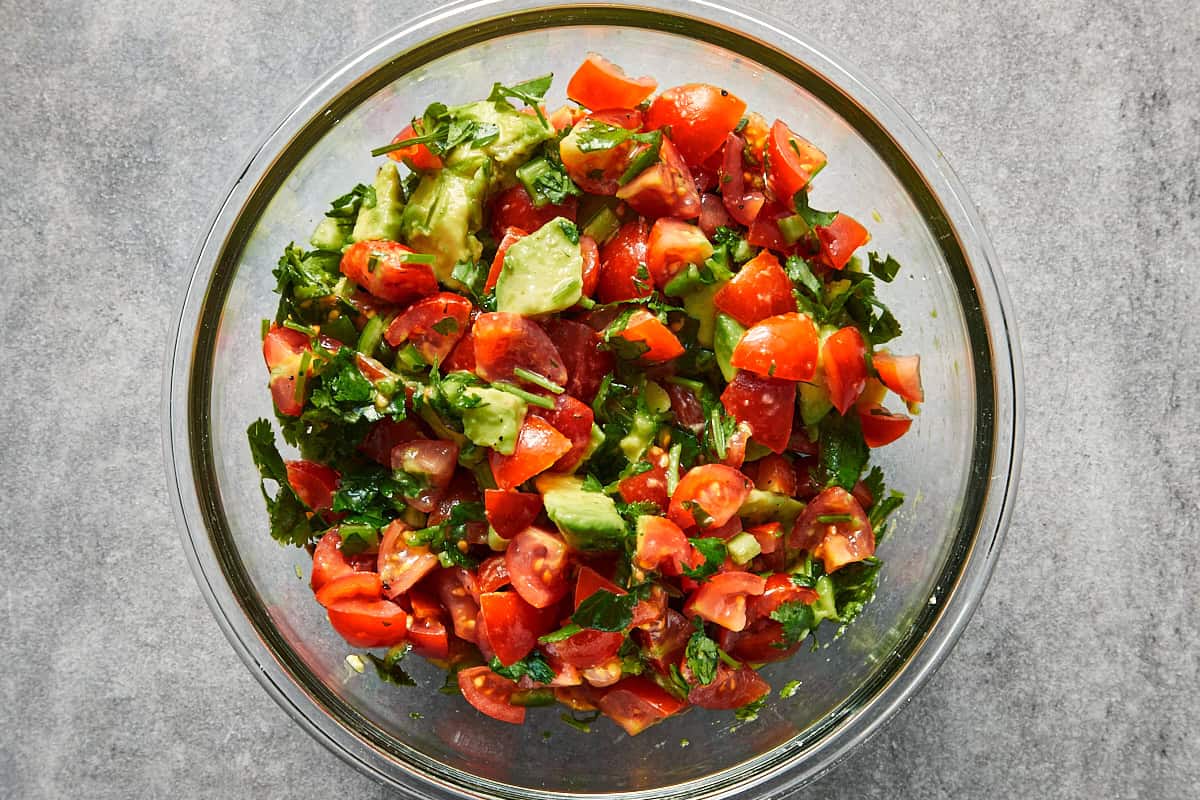 A clear glass bowl filled with fresh diced tomatoes, cubed avocado, chopped cilantro, and jalapenos mixed together.