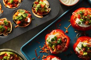 Freshly baked tortilla cups in a muffin tin and stuffed red peppers on a baking sheet, both with melted cheese.