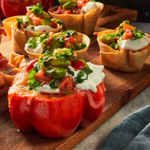 A close-up shot of multiple baked appetizer cups on a wooden serving board, featuring a large, stuffed red capsicum cup in the foreground and several mini tortilla taco cups in the background. All are filled with seasoned chicken, melted mozzarella cheese, and topped generously with sour cream and fresh avocado pico de gallo with coriander.