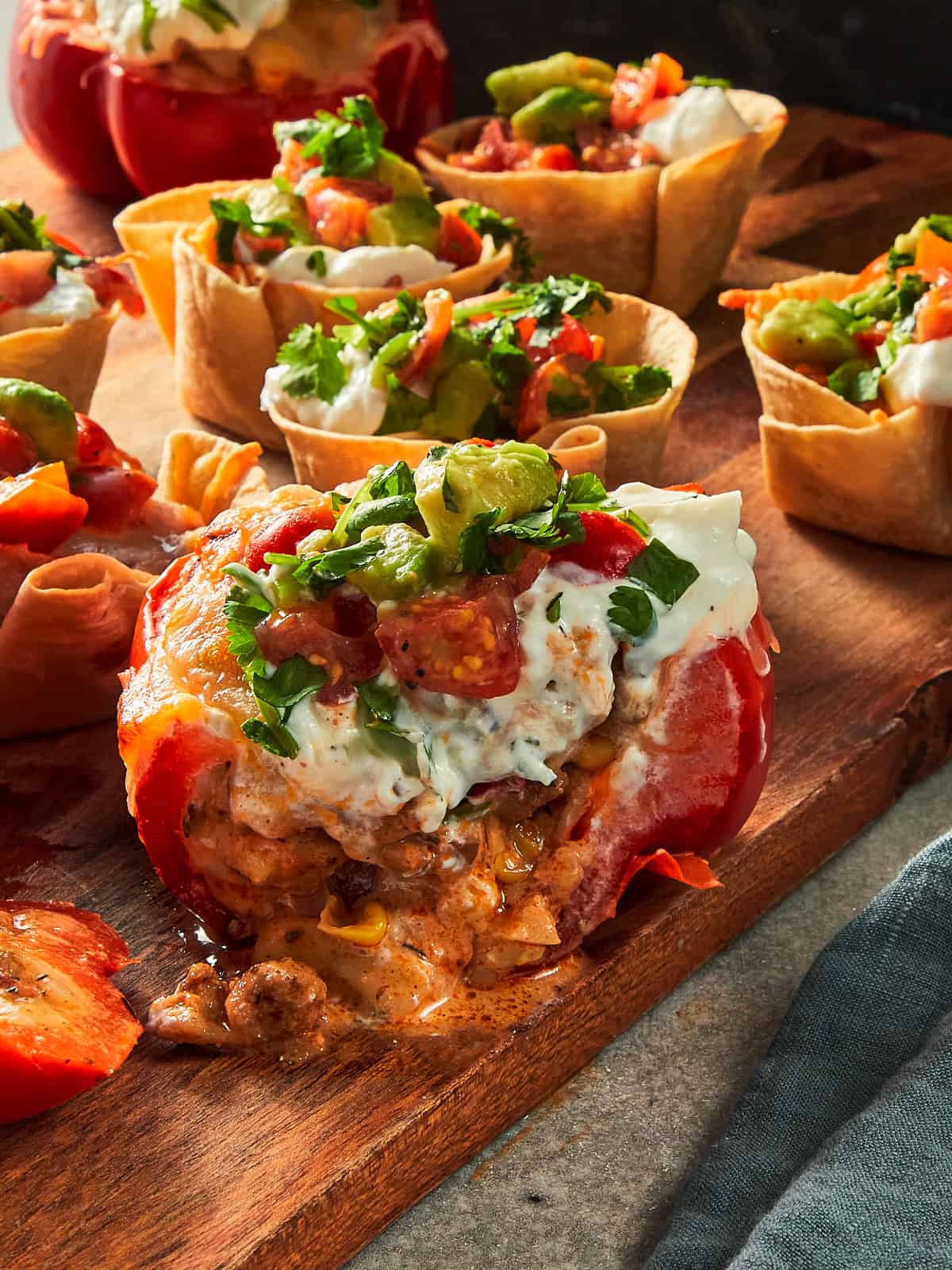 A close-up shot of multiple baked appetizer cups on a wooden serving board, featuring a large, stuffed red capsicum cup in the foreground and several mini tortilla taco cups in the background. All are filled with seasoned chicken, melted mozzarella cheese, and topped generously with sour cream and fresh avocado pico de gallo with coriander.