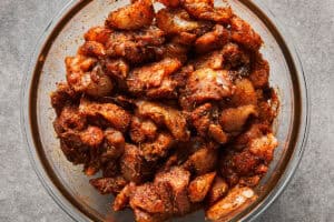 Raw diced chicken thighs coated in red Mexican taco seasoning in a glass bowl.
