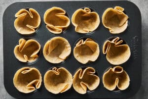 Twelve mini flour tortillas pressed into the holes of a dark metal muffin tin.