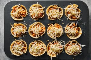 Tortilla cups in a muffin tin filled with chicken mixture and topped with grated cheese.